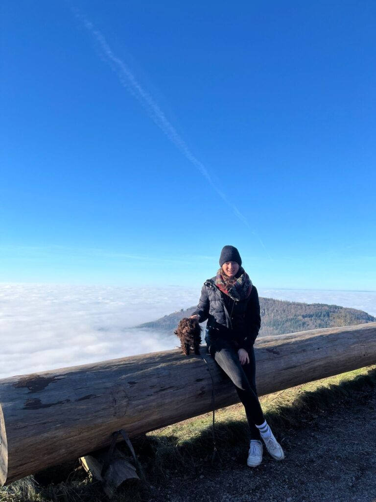 Eine junge Frau und ein Hund lehnen an einem liegenden Baumstamm im Hindergrund sind Berge zu sehen