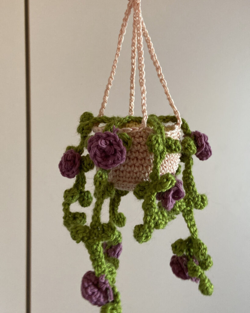 Eine gehäkelte Amigurumi-Hängepflanze in einem kleinen, terracottafarbenen Topf. Aus dem Topf ranken mehrere grüne Blätterstränge herab, die mit kleinen, detailreichen lila Rosenblüten verziert sind.