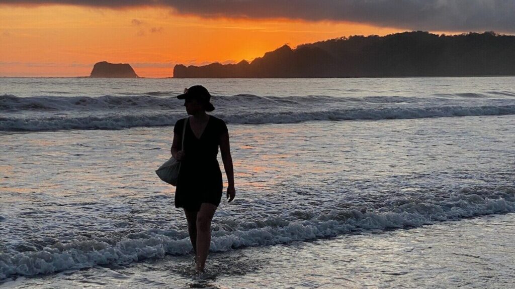 Eine Person läuft am Strand durchs Wasser, während im Hintergrund die Sonne untergeht.
