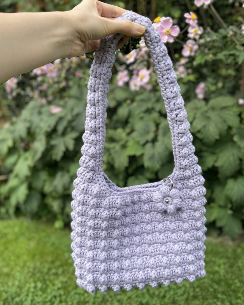 Eine gehäkelte lila Tasche mit Blumen-Anhänger.