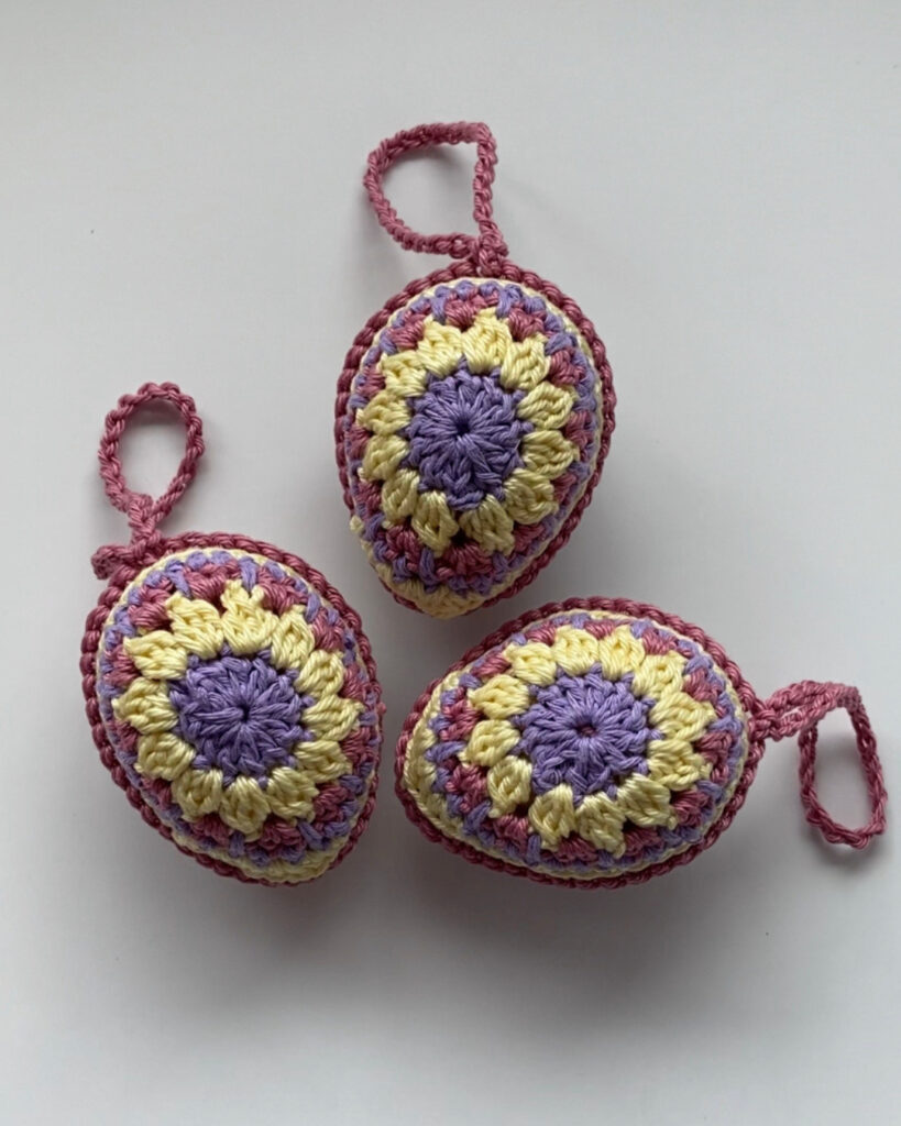 3 gehäkelte Ostereier in den Farben: Lila, Gelb und Magenta in einem Muster gehäkelt.