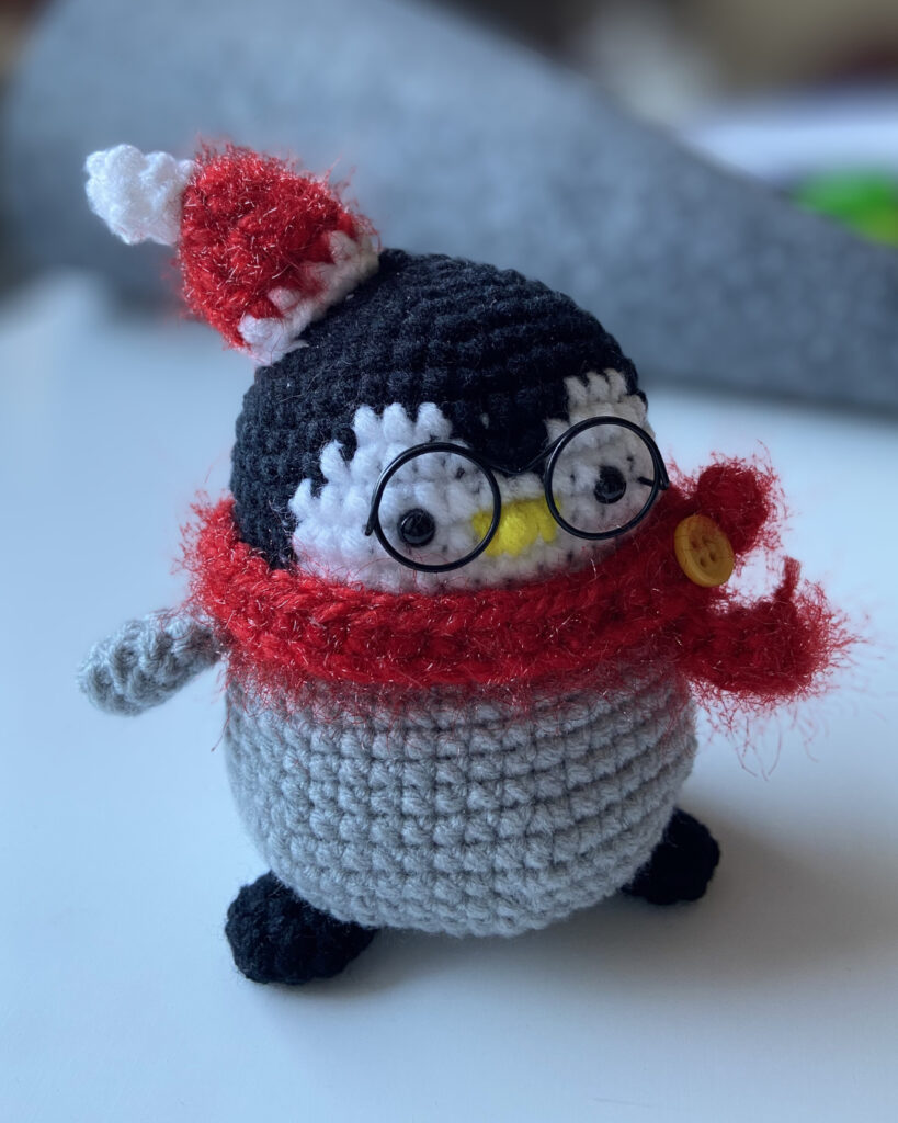 Ein gehäkelter grauer Pinguin im weihnachtlichen Look mit rotem Schal und Weihnachtsmütze und Brille.