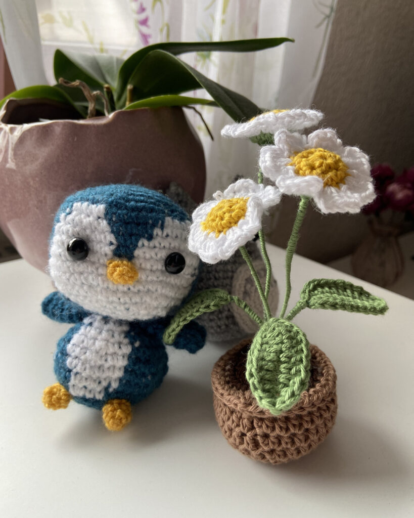 Ein blauer Pinguin (Amigurumi) steht neben einer gehäkelten Topfpflanze.
